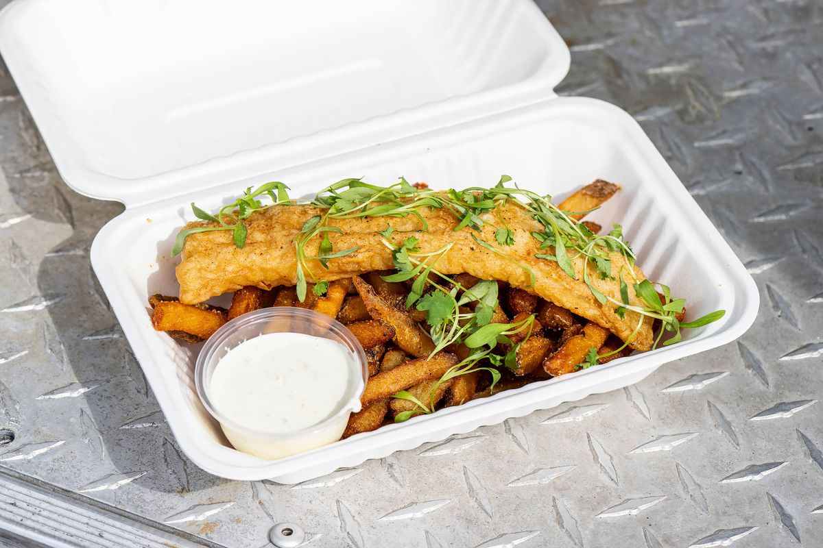 A long piece of fried white fish over a tray of fries from a food truck.