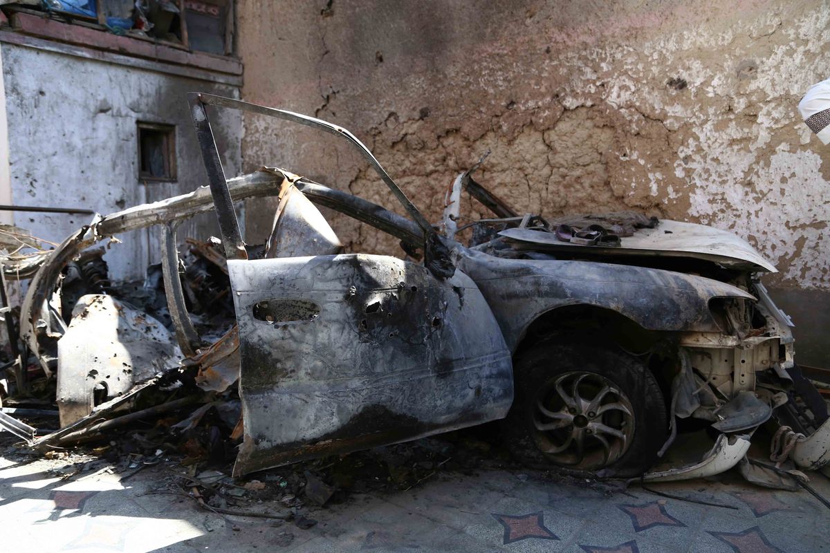 A small car sits in a corner where two buildings meet, crushed and burnt.