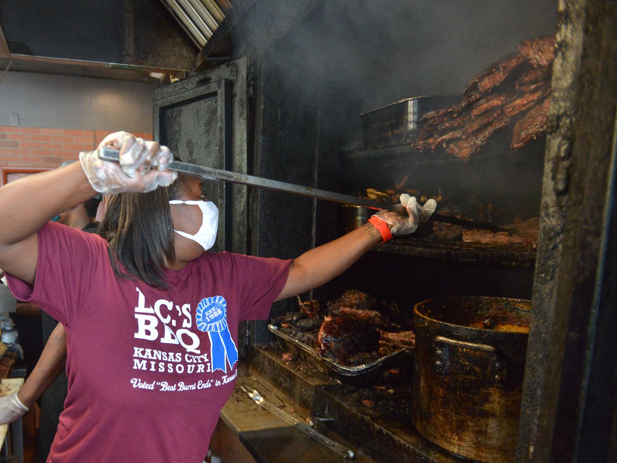 An employee in a branded t-shirt uses a long instrument to adjust meats in a huge oven.