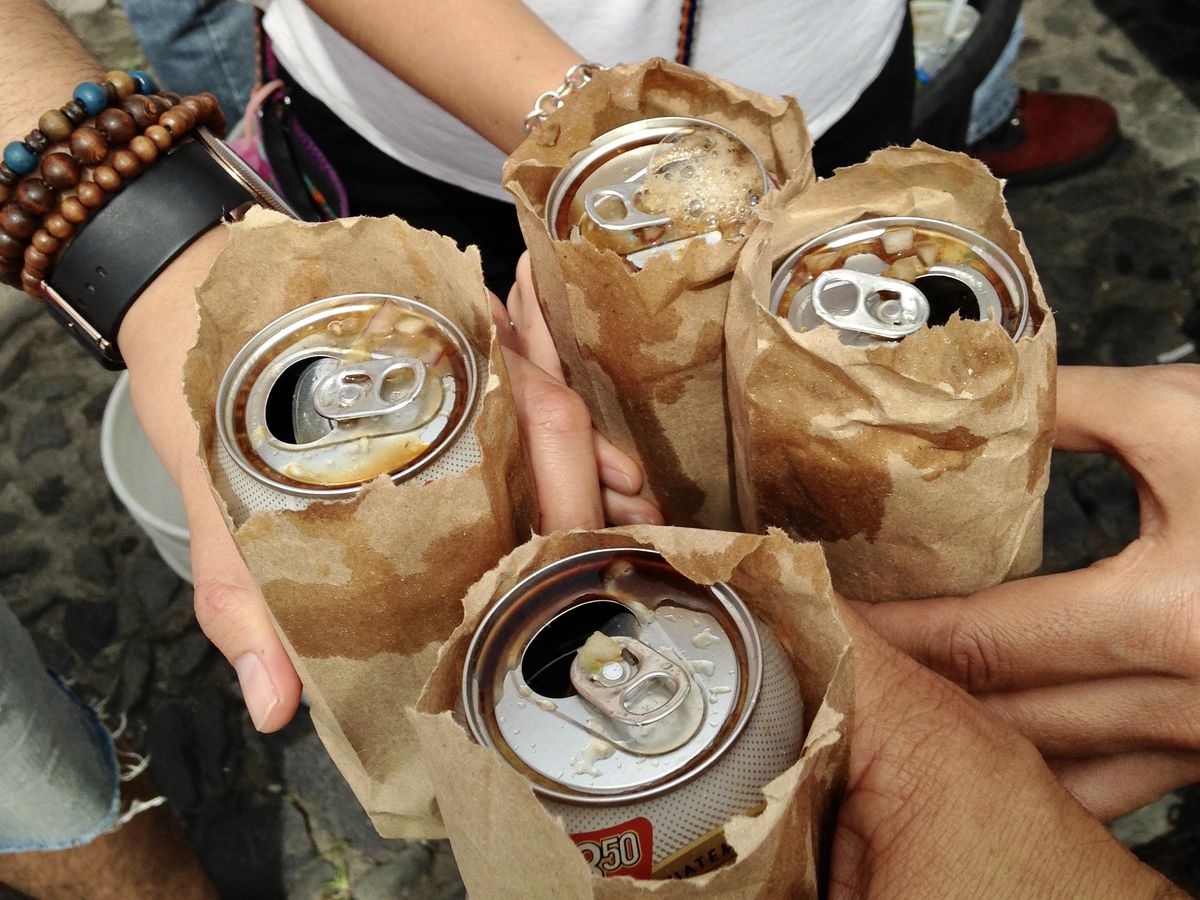 Four hands cheers with beers in brown paper bags topped with tiny bits of salsa. 