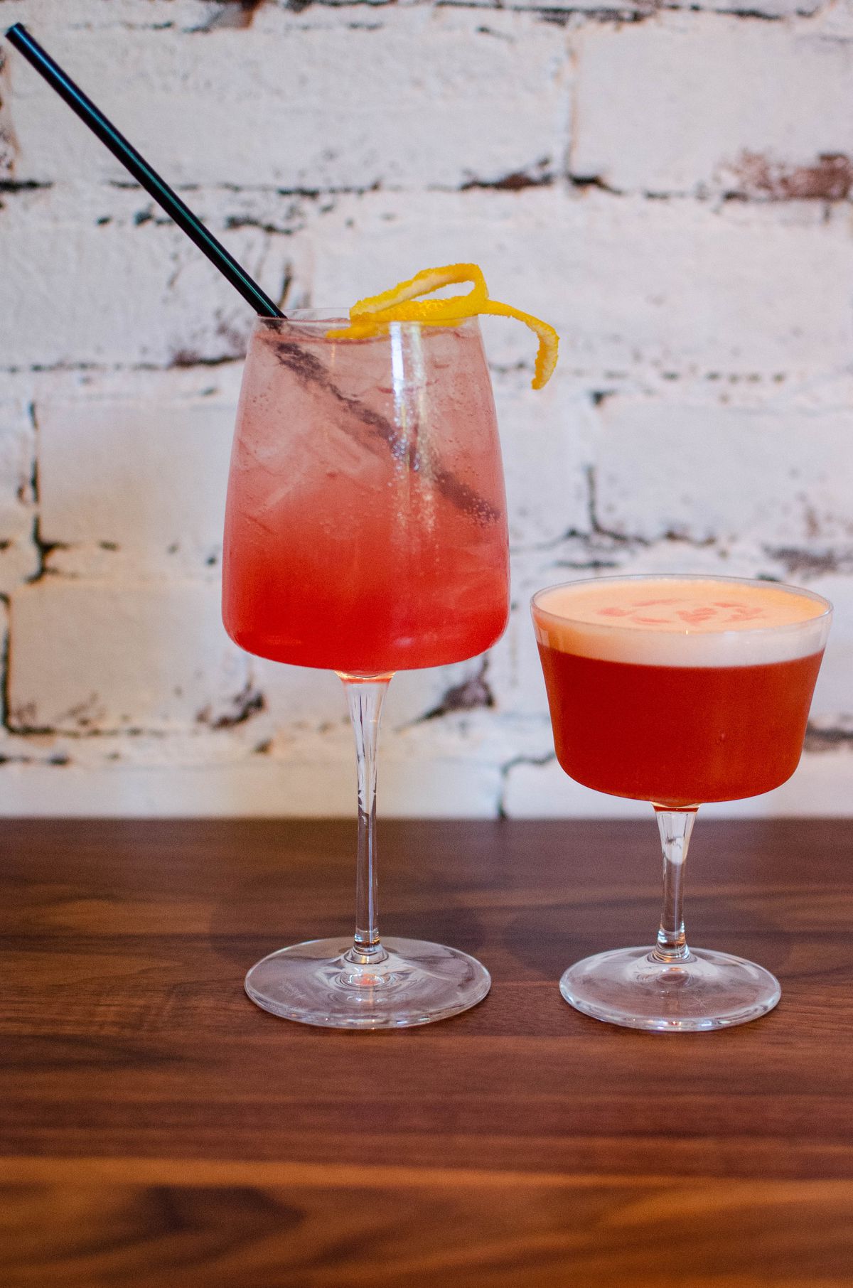 Two pinkish cocktails, one with a foamy top, sit on a wooden table in front of a rustic white brick background.