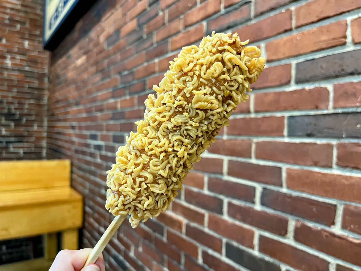 A hand holds up a corn dog on a stick. The corn dog is coated in bits of crunchy, uncooked ramen noodles.