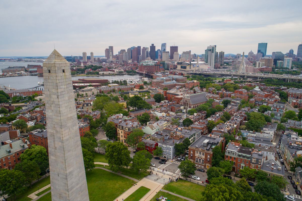 An aerial view of city buildings in Boston. In the foreground is a tall monument. In the distance is a body of water and a skyline with many buildings.