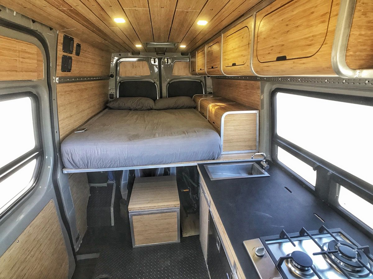 The interior of a van. The walls are wood paneled and there is a bed with grey bed linens. There is a kitchenette with a stove. There are storage compartments under the bed which is elevated.