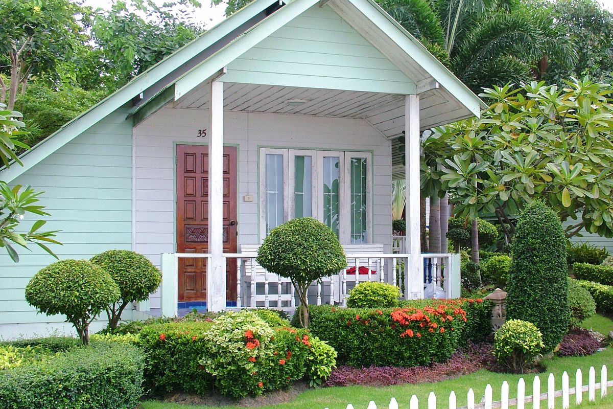 A bungalow style house in a tropical area painted mint green with shrubs and small trees. 