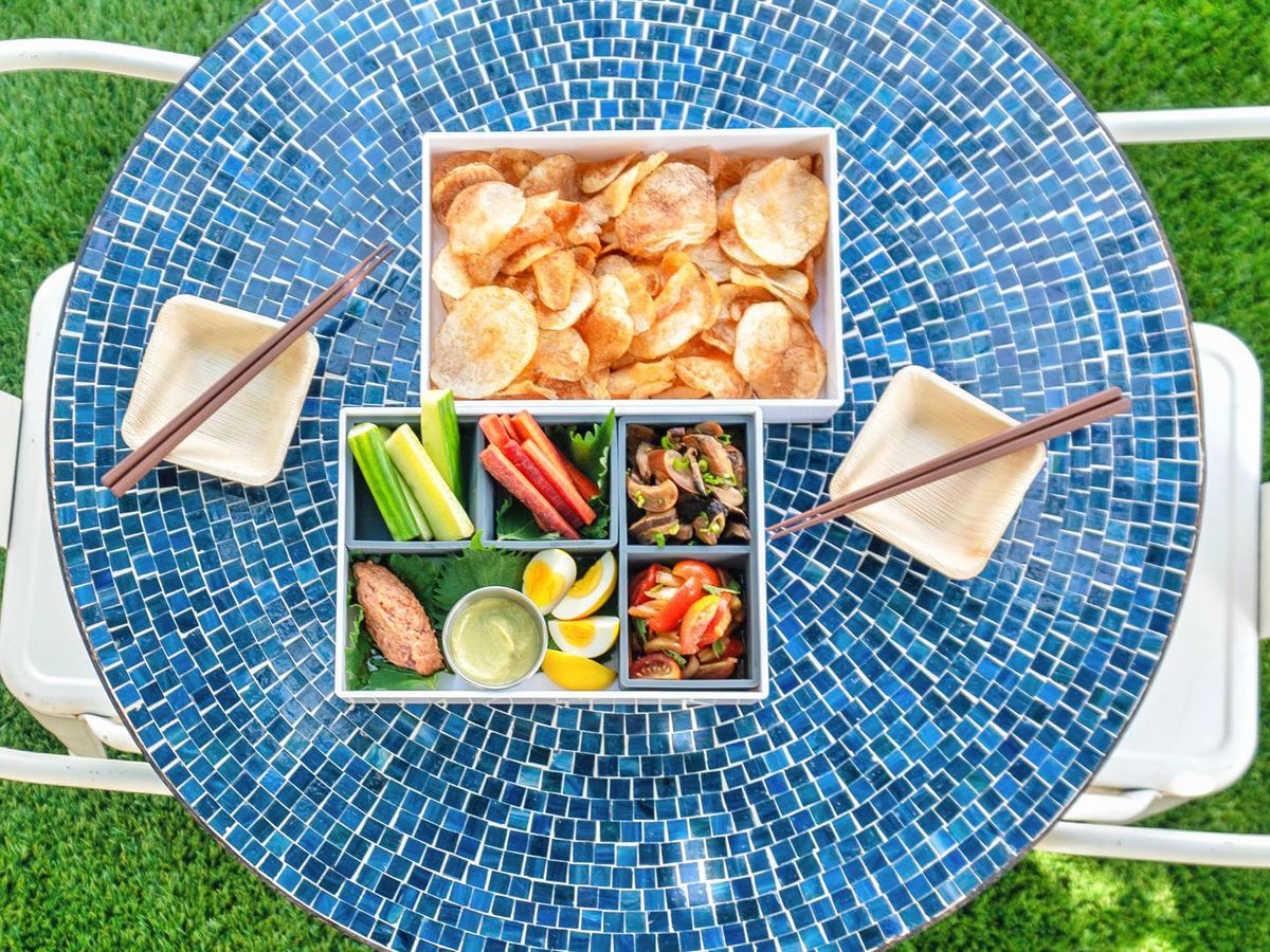 A bento box with chips and another with veggie sticks and other stuff on a blue tile circular table with white plates with chopsticks on them. 