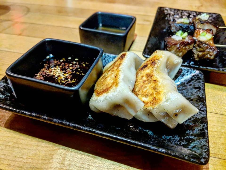 A couple of plump dumplings and a soy dipping sauce sit on a black plate at Momi Nonmi