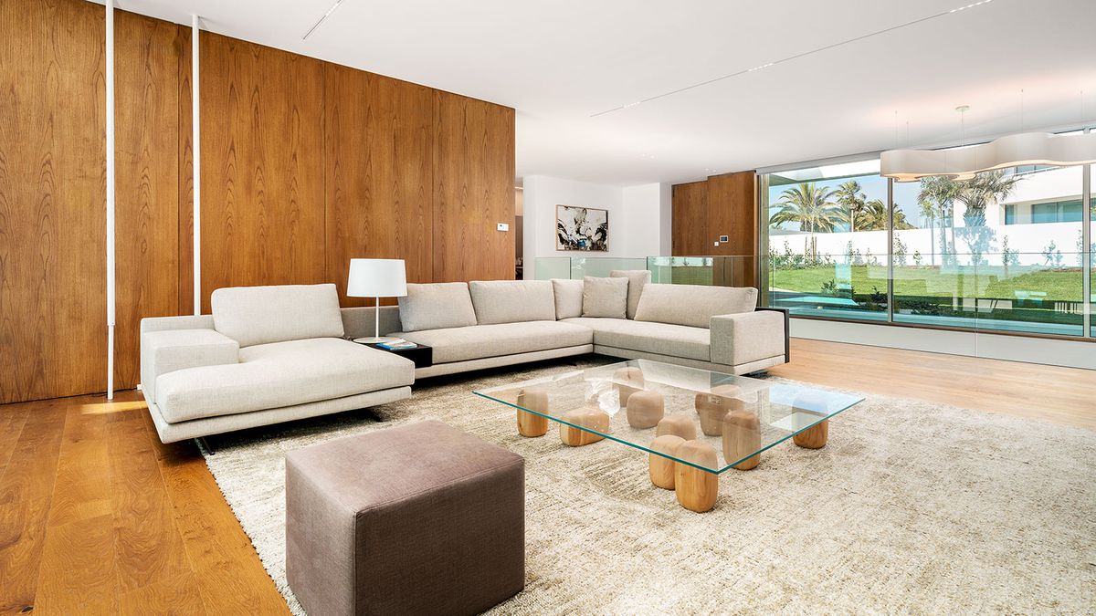A living room with  modern beige sofas and wood paneled walls. A glass coffee table sits on wooden blocks. 