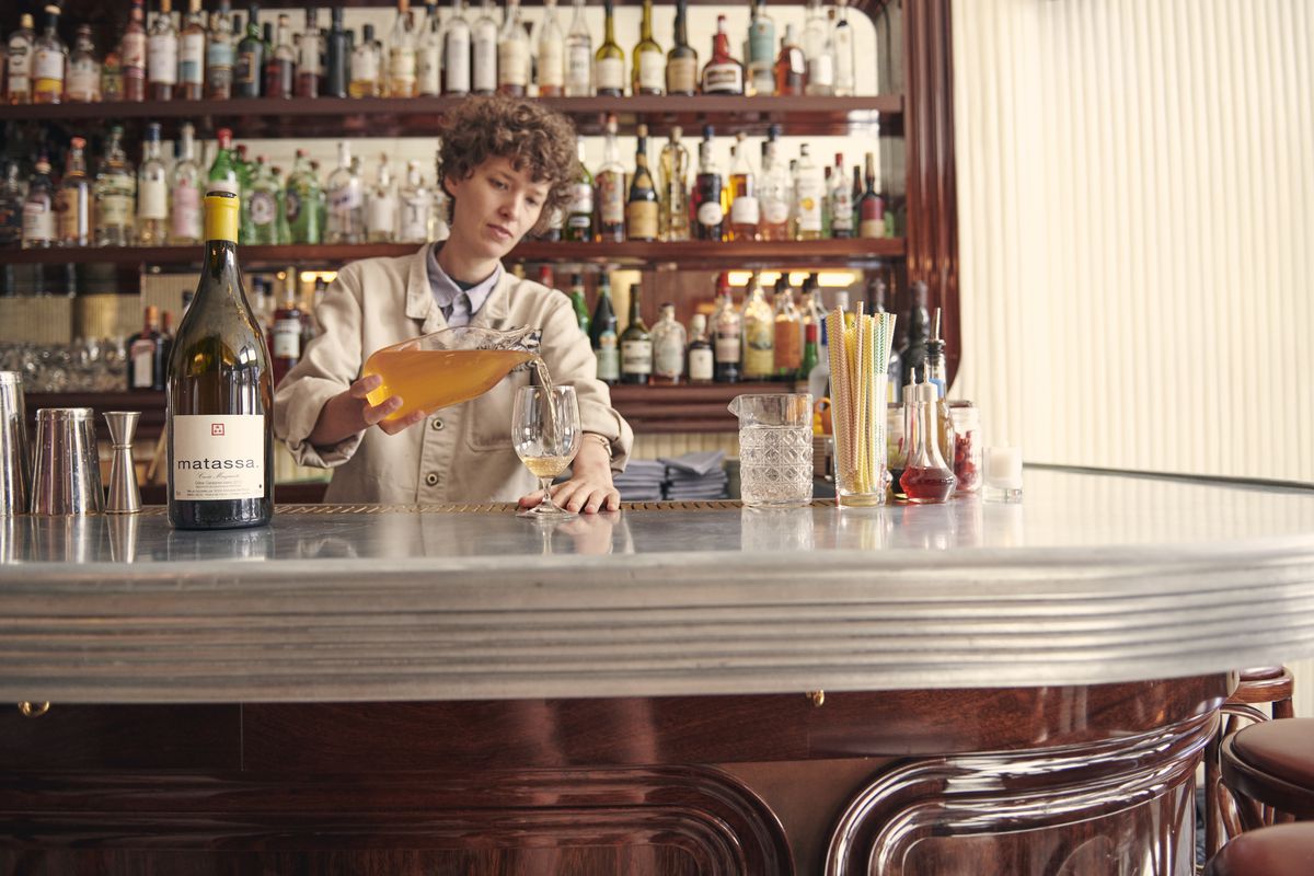 A bartender stands behind a counter, pouring an orange-colored liquid into a wine glass.