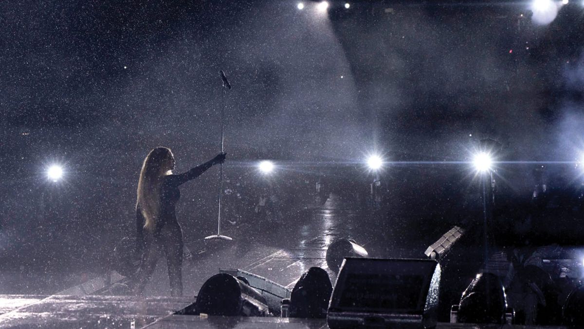 Beyoncé holds a microphone stand up on stage during the Renaissance world tour, as bright lines shine and rain falls.