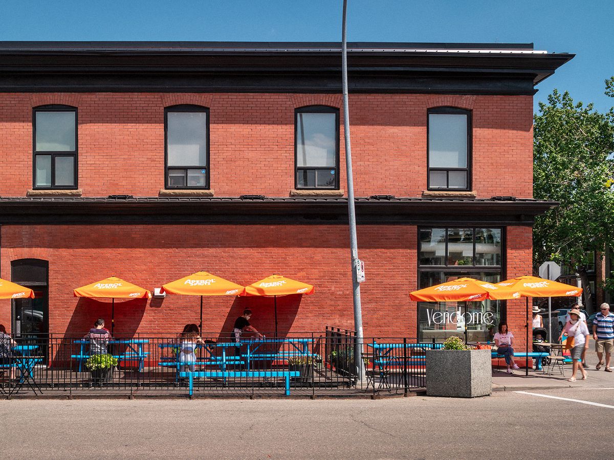 A long red brick building exterior, with a sidewalk patio covered in spots by bright yellow umbrellas, and customers sitting and walking nearby