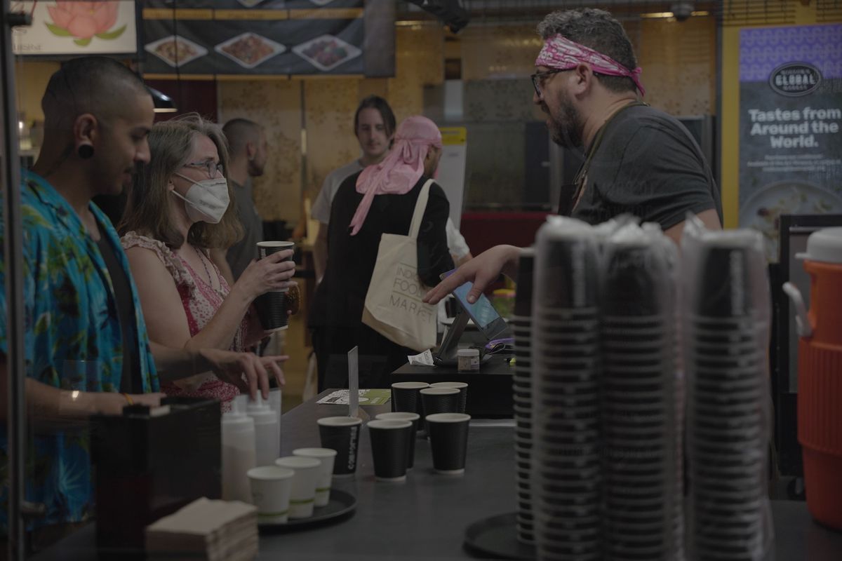 A person wearing a grey shirt, glasses, and a pink bandana talking to a line of people across a counter with paper cups on it. 