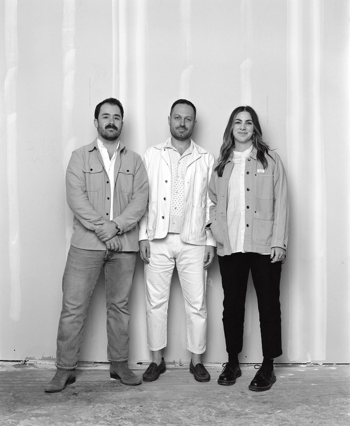 Black and white photo of three people in pants and baggy button-up shirts standing in front of a grey wall with white streaks.