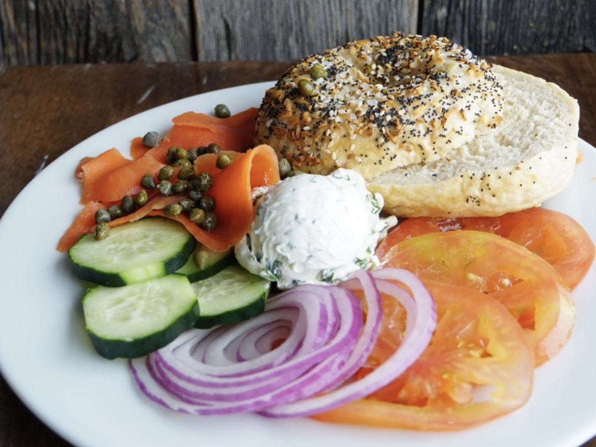 An everything bagel sliced on a plate beside a mound of cream cheese, lox, pickled onions, cucumbers, and tomatoes