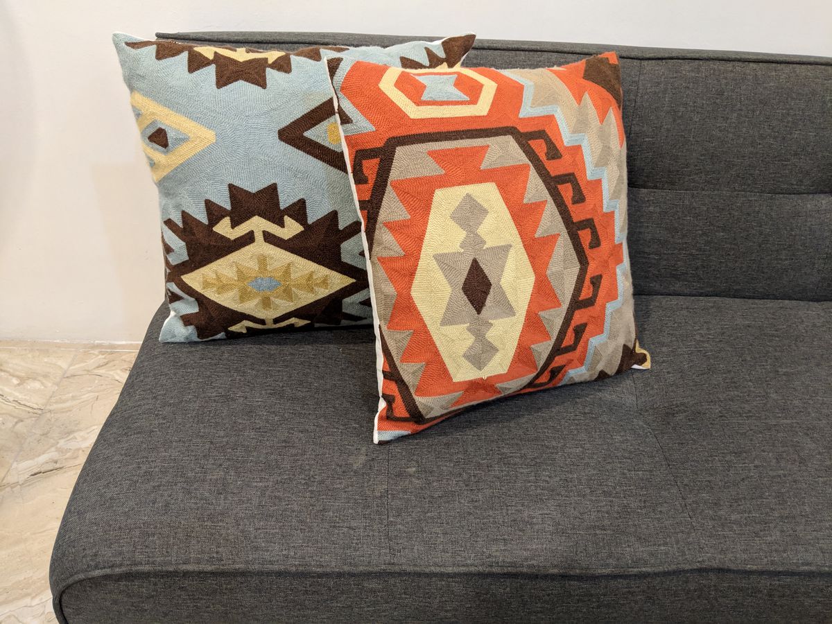 A gray futon with two pillows on it. The pillows have blue, orange, beige, brown, and gray patterns on them.