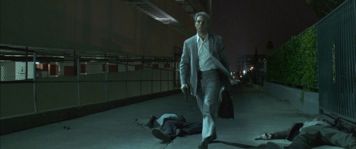 Tom Cruise, holding a gun and a bag, walks past some dead bodies after some murdering in Collateral.