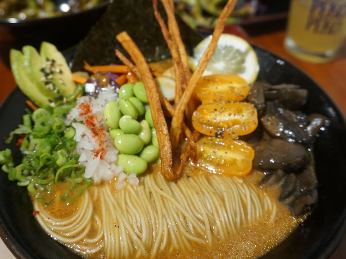 A bowl of ramen, with noodles curled to one side in the broth. Toppings include edamame, scallion, onion, tomato, mushrooms, and some sort of crunchy sticks