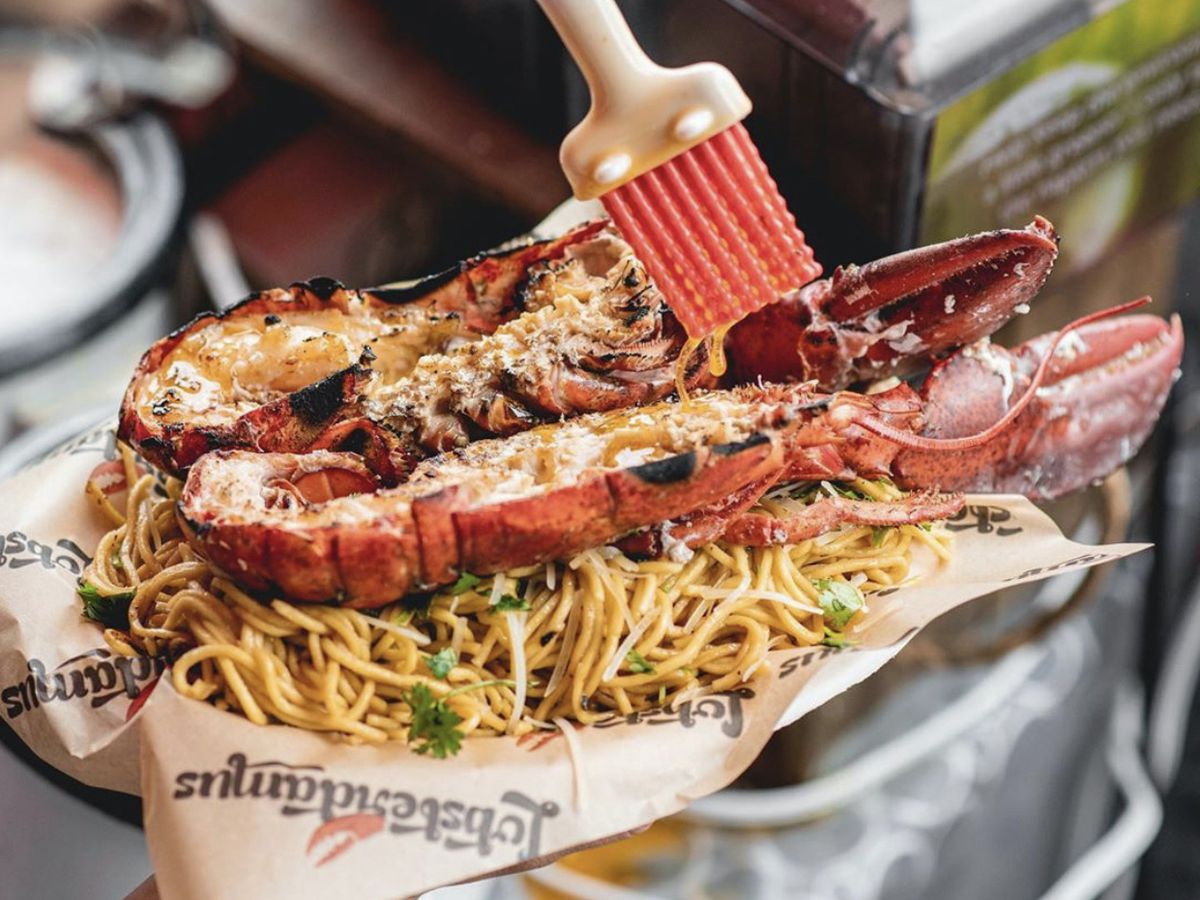 A cut-open lobster being basted with butter, set on top of noodles