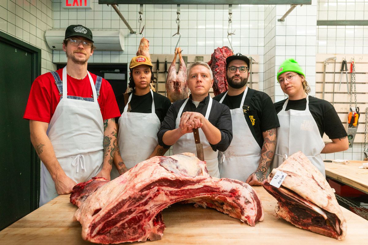 Several butchers stand around a table with a quarter of meat. 