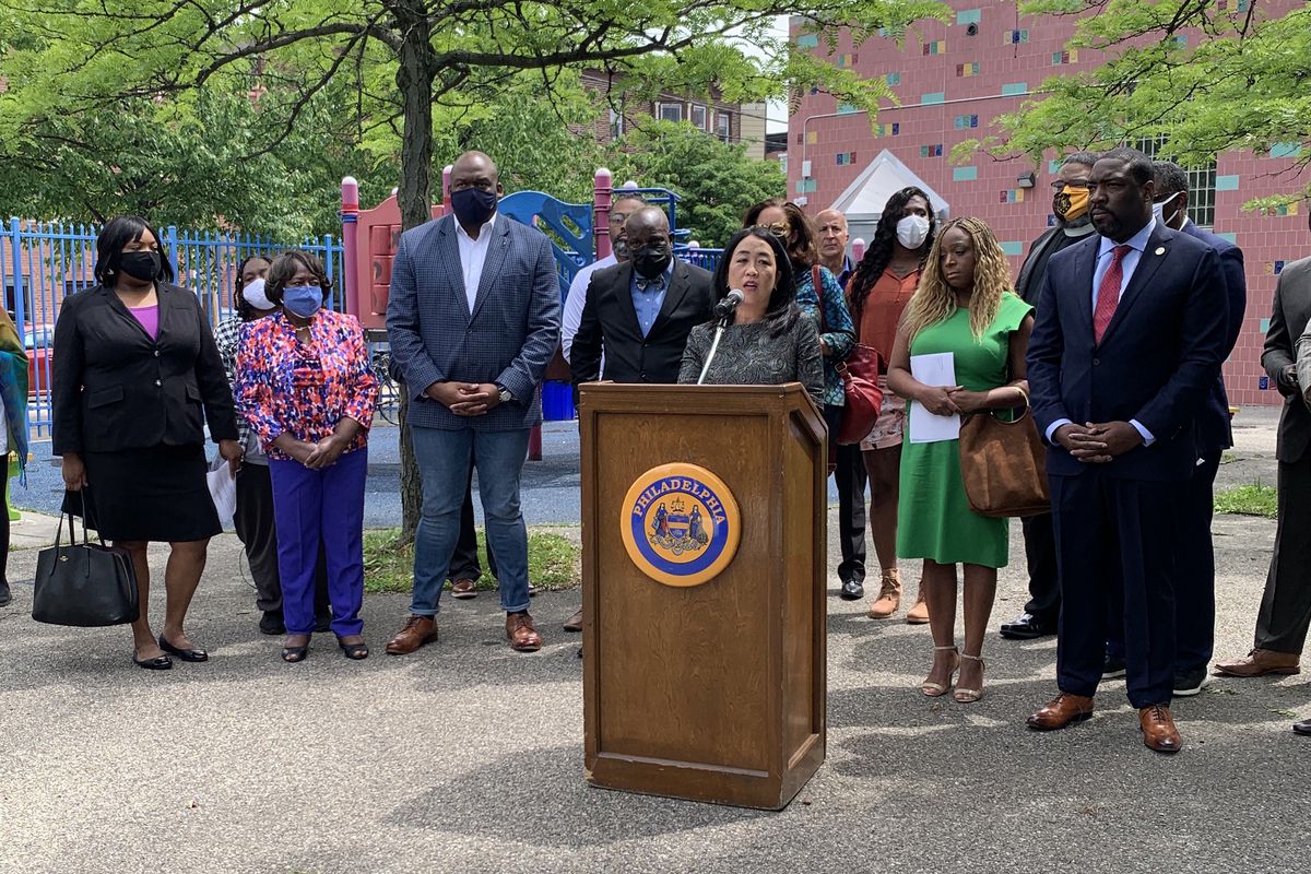 Council member Helen Gym is standing at a lectern outside and is surrounded&nbsp;by several other council members, community and religious leaders.