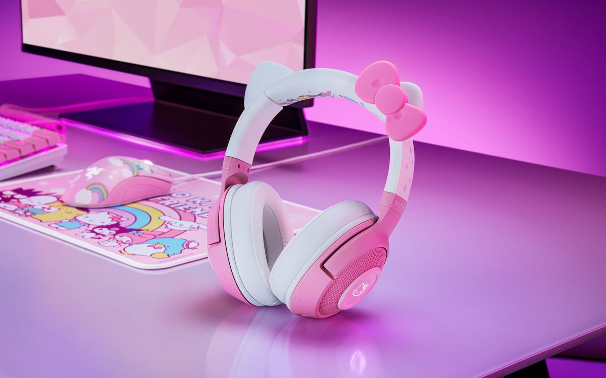 A pair of Hello Kitty headphones from Razer sitting on a desk with a gaming computer setup in the background