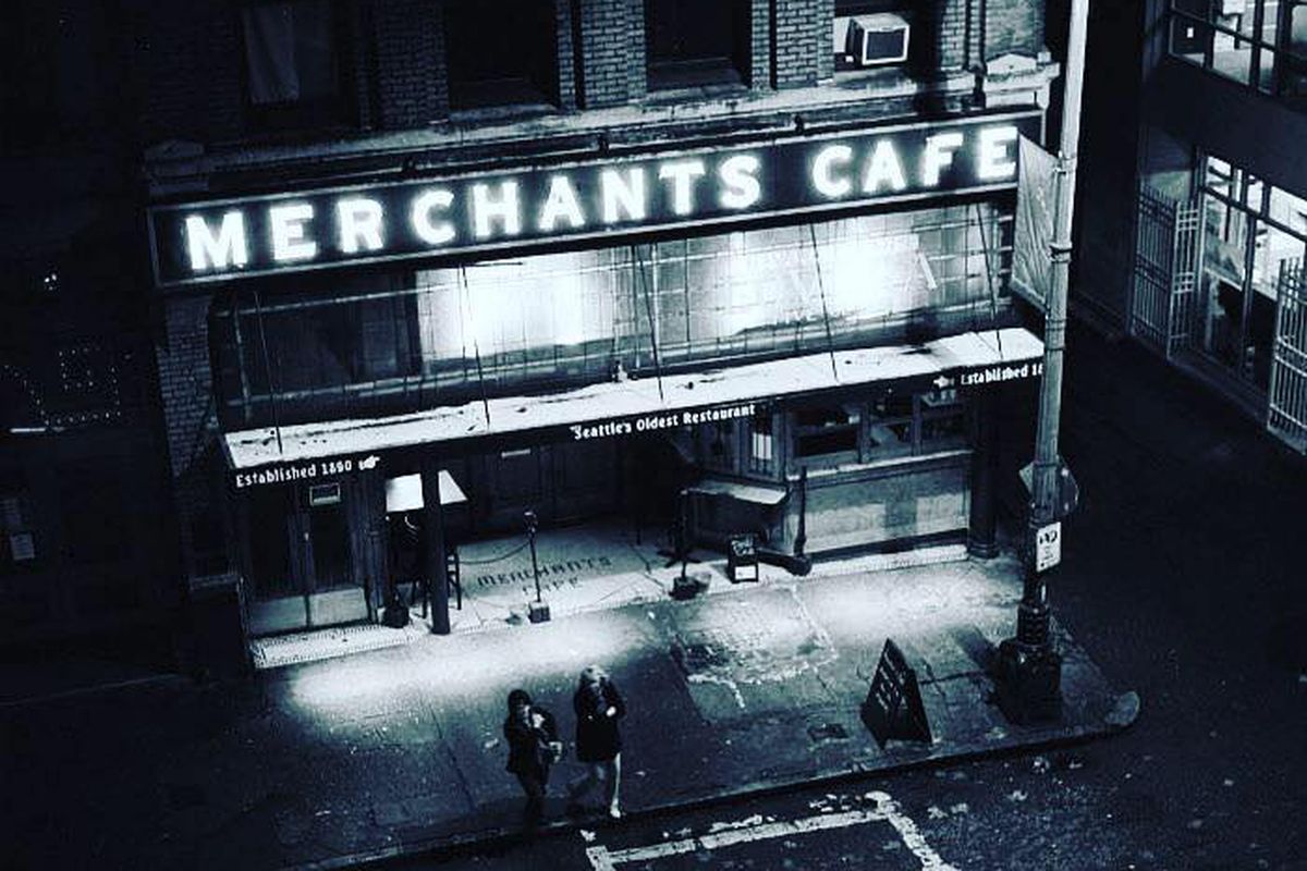 A black-and-white photo of the exterior of Merchant’s Cafe, with the restaurant’s iconic sign lit up at night.