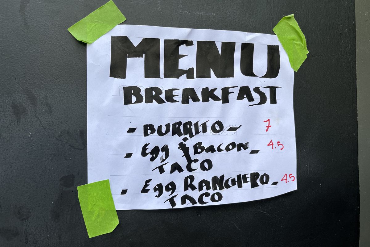 A white sheet of paper taped to a wall with green tape. It has the word MENU written in large capitals, followed by Breakfast, followed by Burrito; Egg bacon taco; and egg ranchero taco.