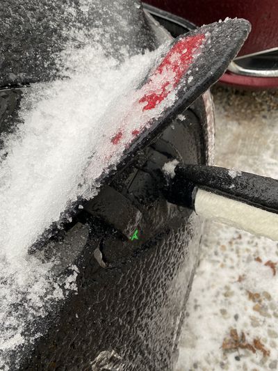 This is a closeup of the charge port door of a Model 3, with the Tesla T logo lighting up green and a J1772 plus Tesla adapter plugged in. There is snow and ice stuck around the whole port and door.