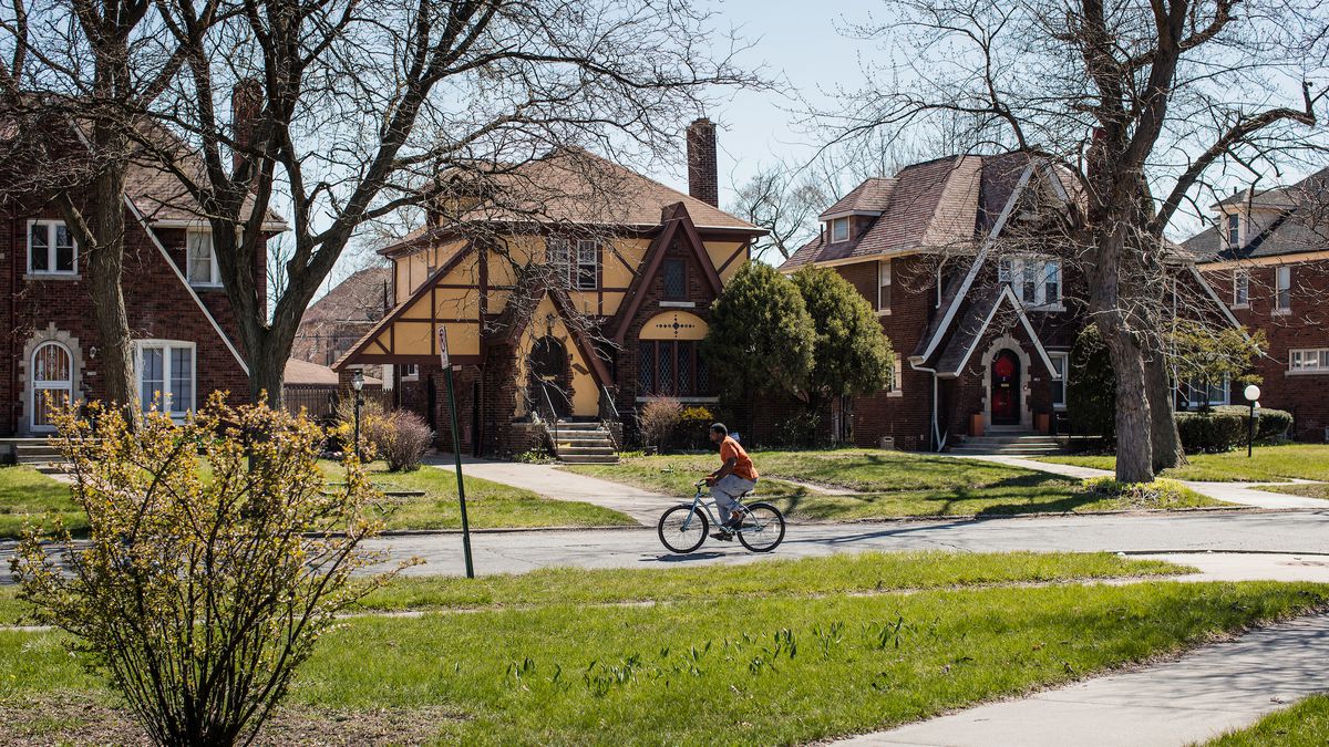 A neighborhood street, with three tudor style houses, a man on a bike is riding by. 