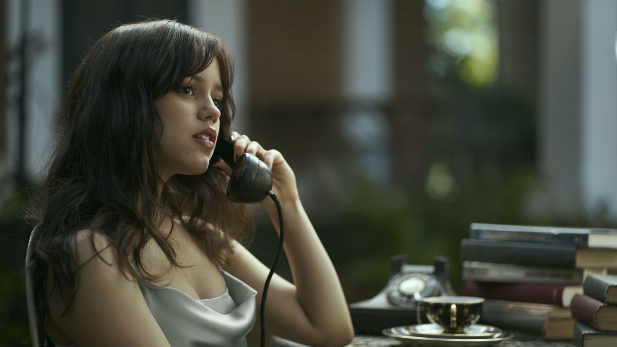 Cairo Sweet (Jenna Ortega, in a silver silk dress baring her arms and cleavage) sits at a table covered with books talking on an old-fashioned dial telephone in Miller’s Girl