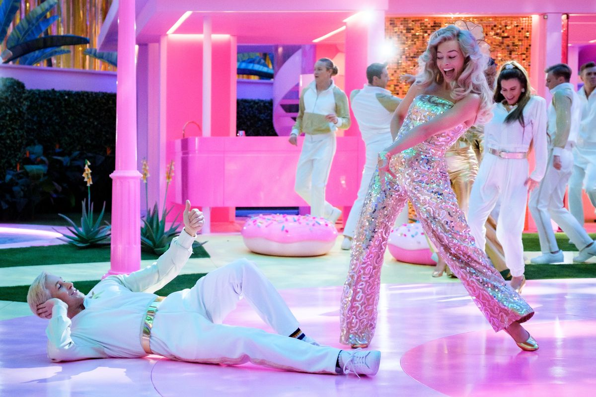 Barbie (Margot Robbie), in a silver spangly jumpsuit, grins at Ken (Ryan Gosling), in an all-white outfit, lying on a shiny pink dance floor with other dancing Barbies and Kens in the background in Greta Gerwig’s live-action 2023 movie Barbie