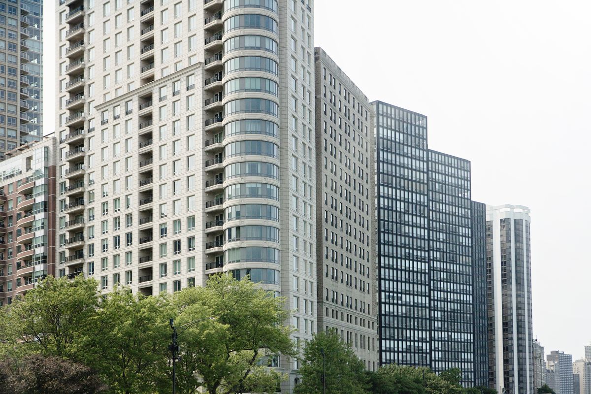 Tall towers with beige and black exteriors and tiny windows face the lakefront. Trees are planted in front of the structures.