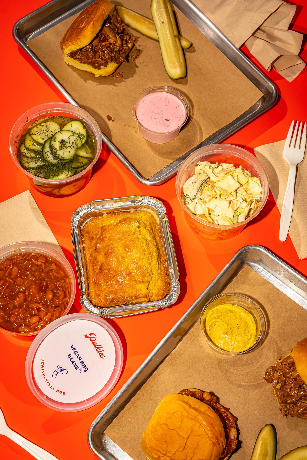 Several open containers of food are placed on an orange sheet including sandwiches, pickles, and salad