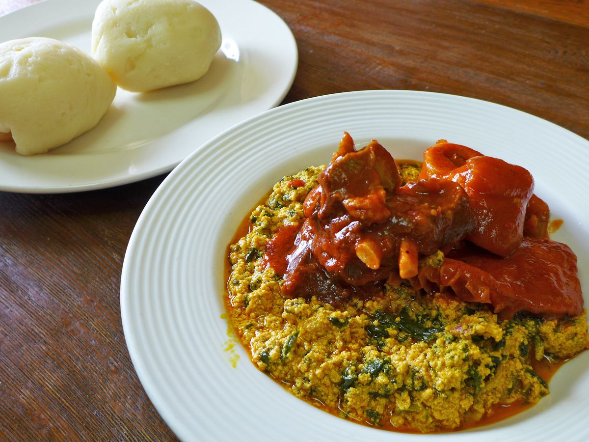 White plates with egusi and fufu are lain out on a wooden counter.