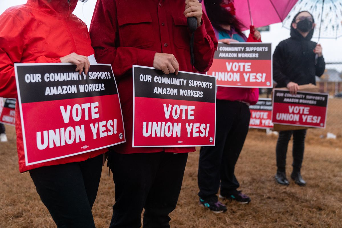 A line of protesters stand outside, holding red signs reading “Vote Union Yes!”