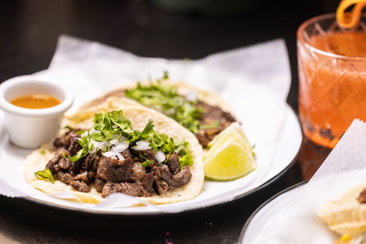 A pair of carne asada tacos are serve on a metal tray with a sheet of white paper on top, and accented with a wedge of lime and a small tray of sauce. A glass of orange agua fresca sits to the right.