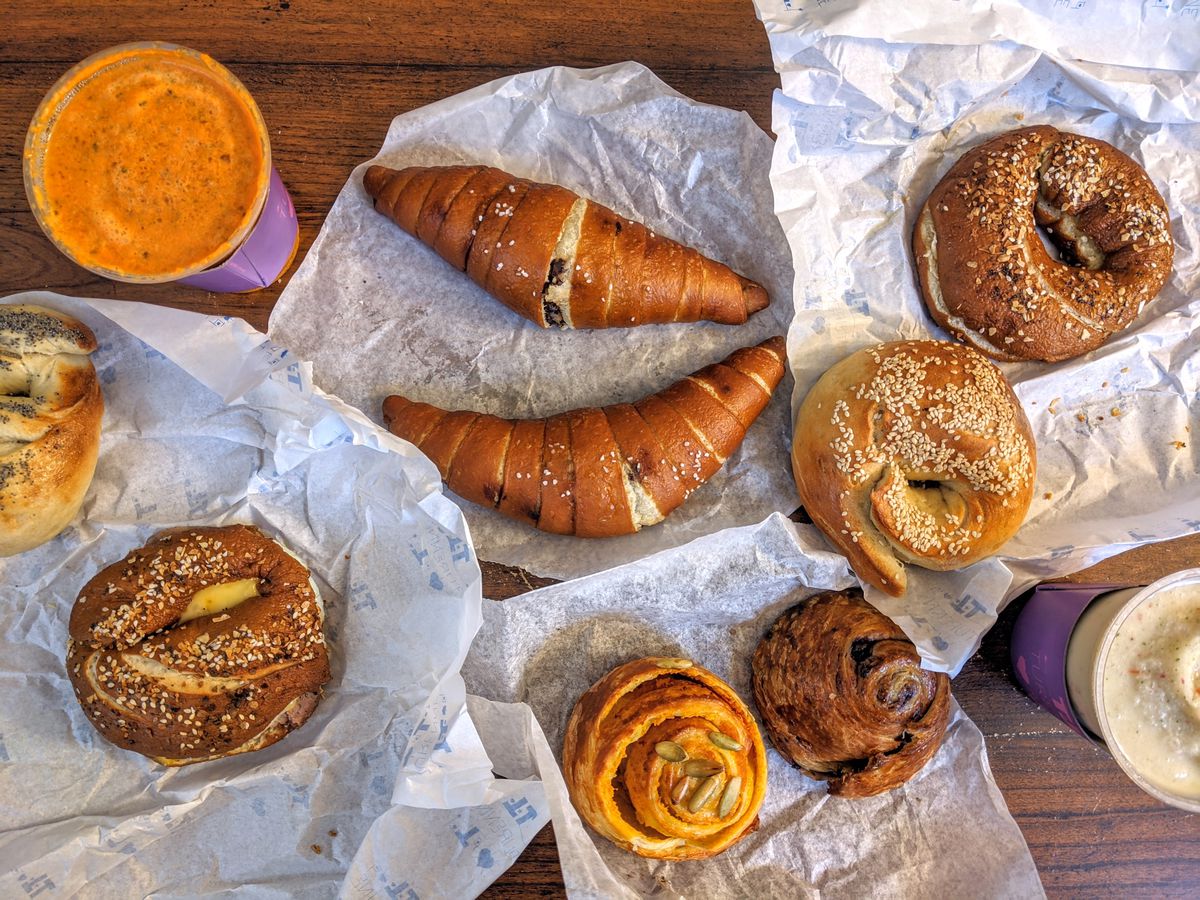 Overhead view of food spread out on white paper over a wooden tabletop: bagels, croissants, smoothies, and more.