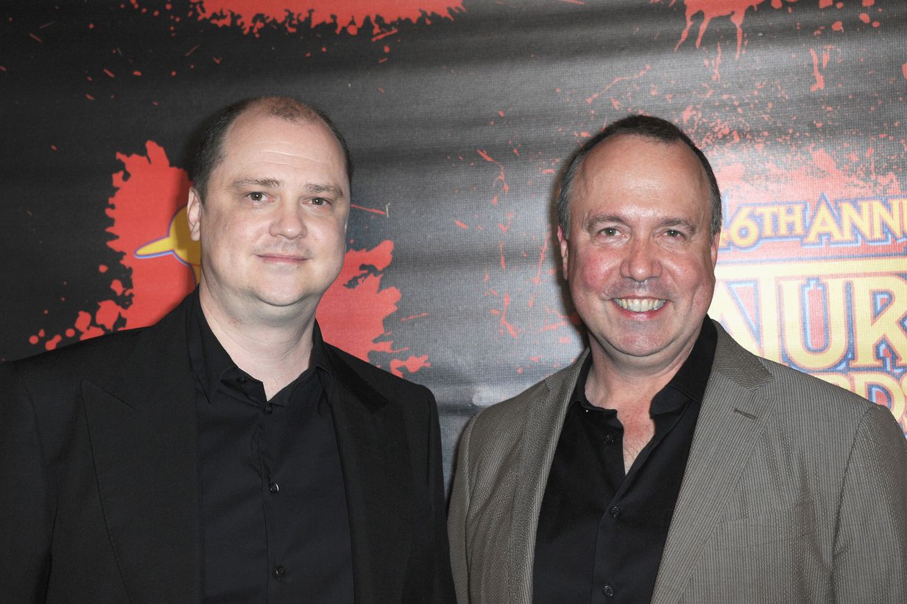 Two men in black button down shirts and dark blazers — one black and the other gray — standing in front of a Saturn Awards backdrop.