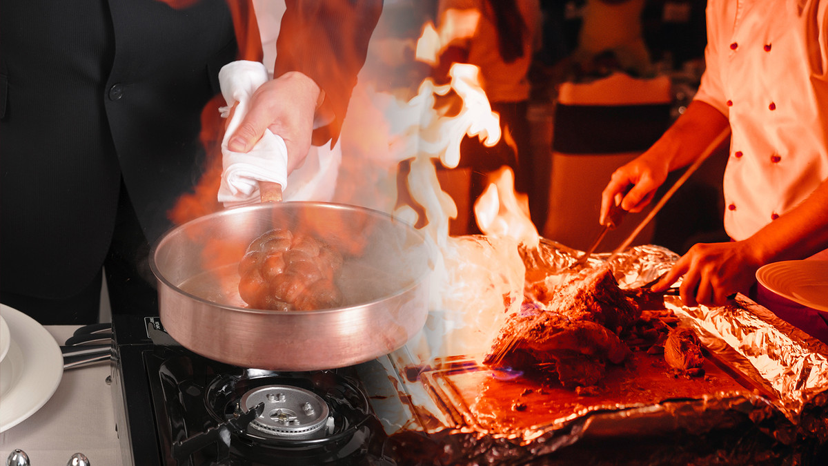 A flaming pan next to a server in a chefs coat cutting meat.