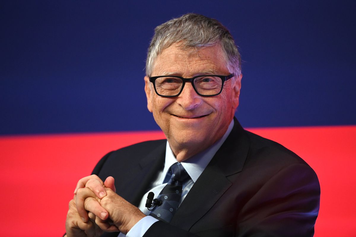 Bill Gates smiling and sitting onstage in front of a red and blue background.