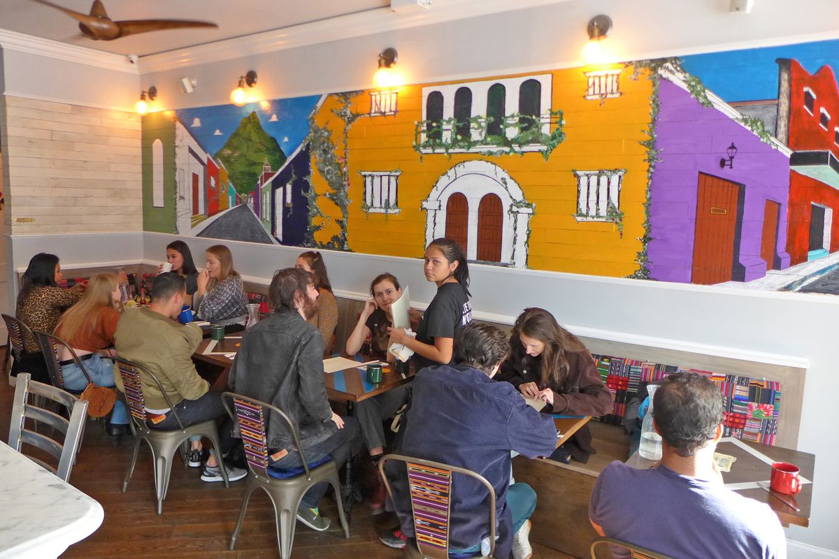 A few tables with diners in jeans stand before a wildly colorful mural of a Guatemalan village.