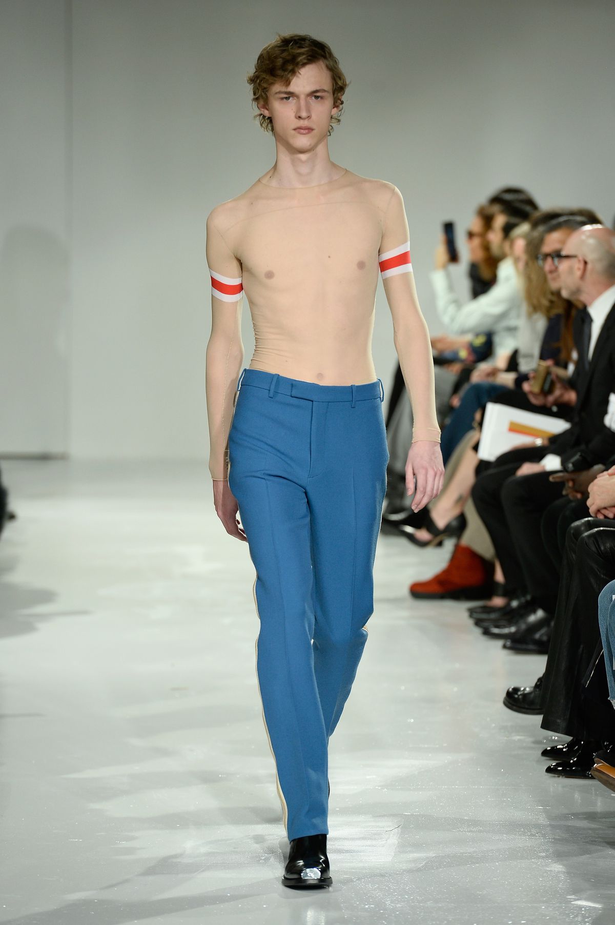 A model wears blue trousers and a transparent shirt with red and white arm bands.