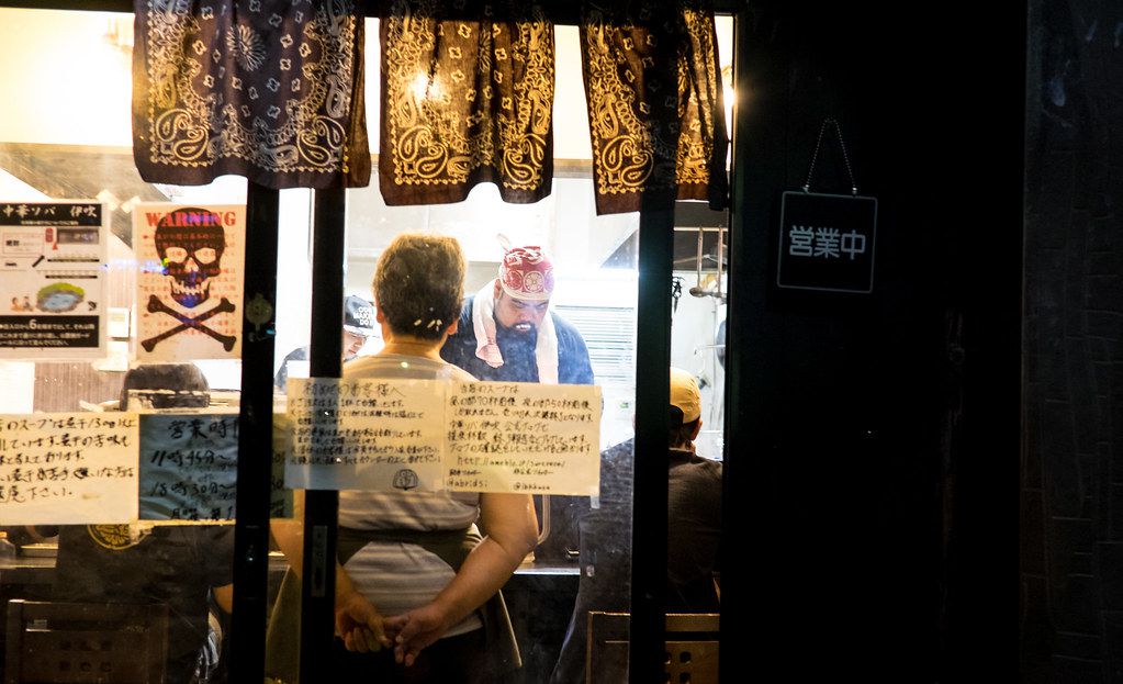 Through a darkened window, a chef working behind a ramen counter.