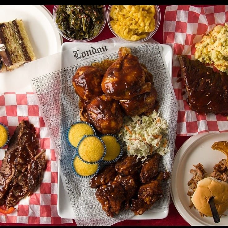From above, a table with lots of dishes lined with a checkerboard tablecloth. dishes include barbecued chicken, muffins, coleslaw, collard greens, mac and cheese, cake, sandwiches, and other barbecued meats
