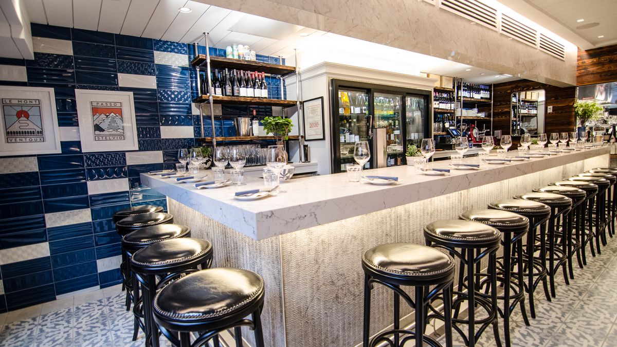 Shiny black stools line a white marble bar. The floor has Spanish-style tiles, and there are dark blue, glossy tiles on the wall.