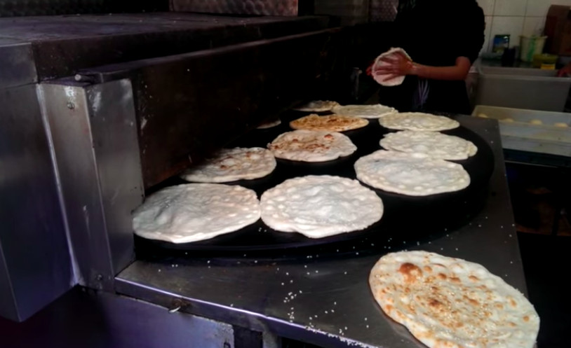 A grill covered in discs of uncooked flatbread dough, ready to go into an oven, with a baker in the background shaping one of the breads