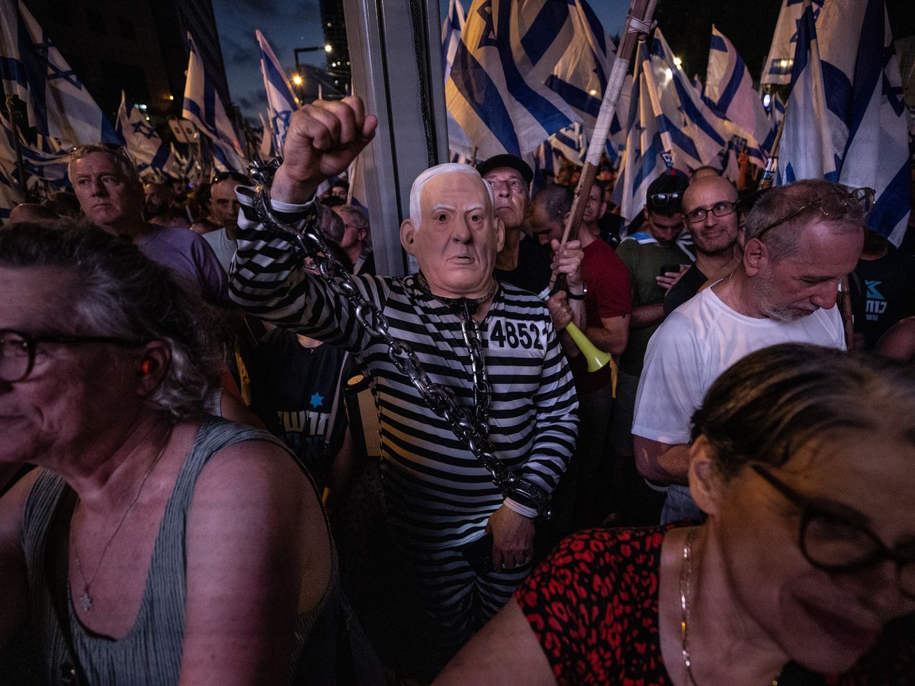 A crowd of protesters, waving blue and white Israeli flags, march through Jerusalem, Israel, on July 23, 2023. One protester wears a black and white striped inmate costume and a rubber mask resembling Netanyahu’s face, and raises a fist in the air.
