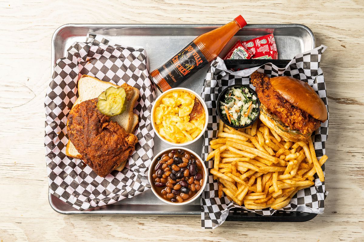 A metal tray holds several dishes including fried chicken and pickles on white bread, beans, hot sauce, french fries, and more.