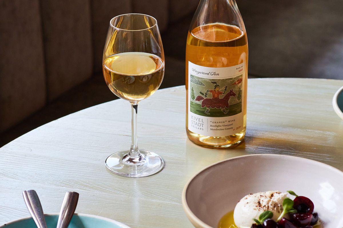 A blonde wood table with a bottle of rosé wine and glass with a bowl of cheese and a blue plate.
