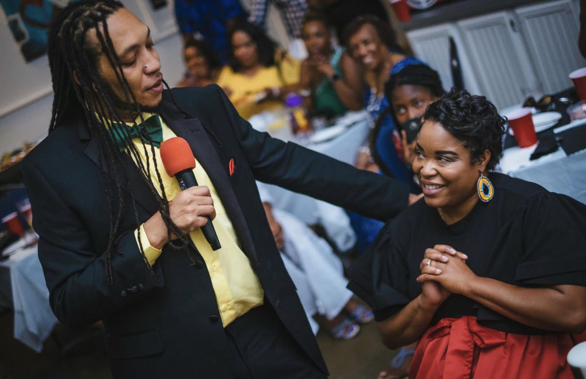 A Black man with long dreadlocks speaks into a microphone and gestures to a Black woman, who is smiling.